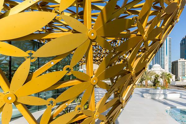 An dem lichtdurchlässigen Dreiecks-Gitterwerk aus verschweißten Stahlprofilen ist an jedem Knotenpunkt eine goldene Blüte angebracht.